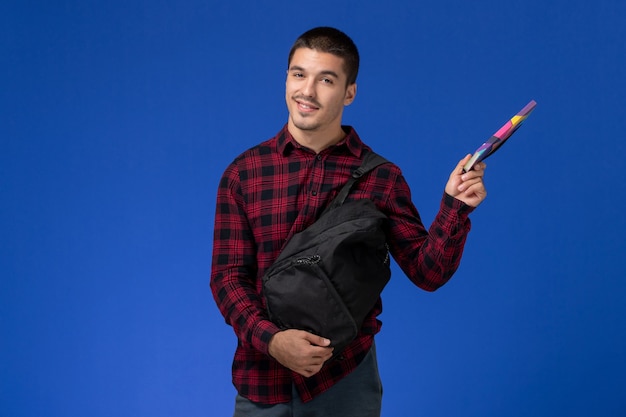 Front view of male student in red checkered shirt with backpack holding copybook on the blue wall