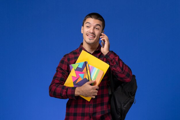 파란색 벽에 전화 통화 카피 북 및 파일을 들고 배낭 빨간색 체크 무늬 셔츠에 남성 학생의 전면보기