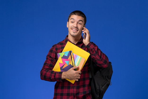 コピーブックと青い壁に電話で話しているファイルを保持しているバックパックと赤い市松模様のシャツの男子学生の正面図