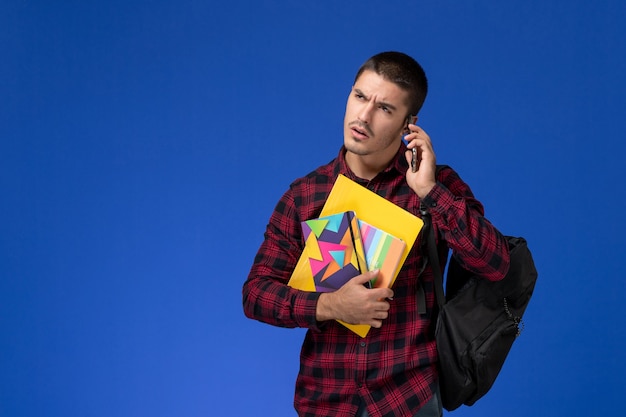 Front view of male student in red checkered shirt with backpack holding copybook and files talking on the phone