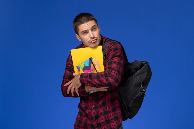 Front view of male student in red checkered shirt with backpack holding copybook and files with phone on the blue wall