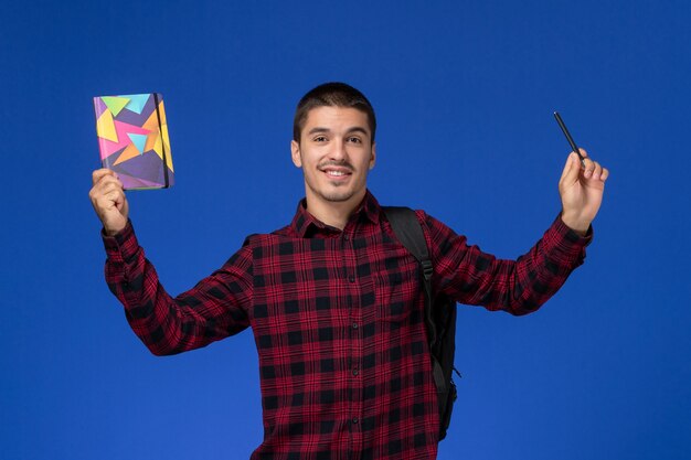 Front view of male student in red checkered shirt with backpack holding copybook and pen on light-blue wall