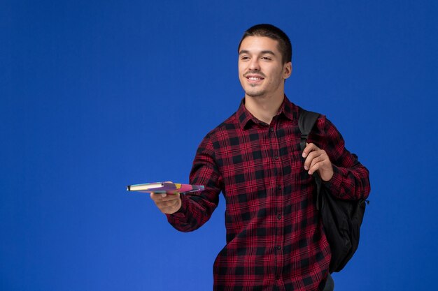 Front view of male student in red checkered shirt with backpack holding copybook smiling on light blue wall