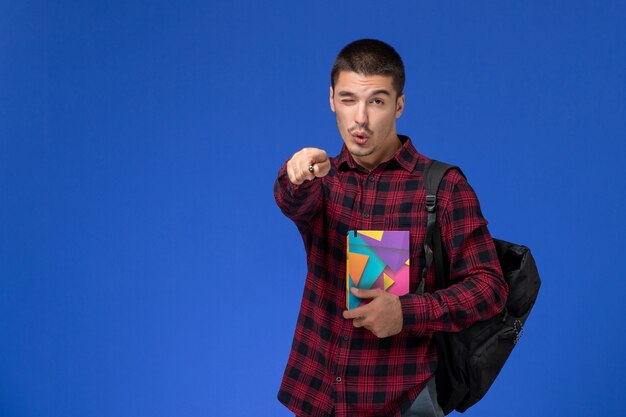 Front view of male student in red checkered shirt with backpack holding copybook and winking on light-blue wall