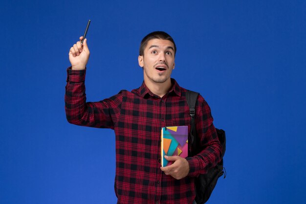 Front view of male student in red checkered shirt with backpack holding copybook