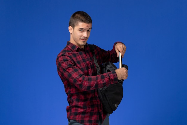 Front view of male student in red checkered shirt with backpack holding copybook