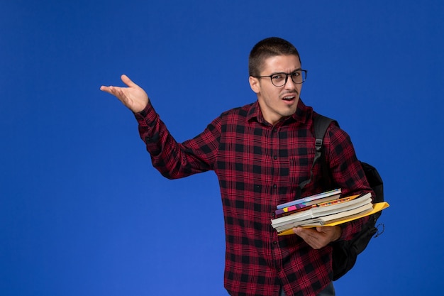 Front view of male student in red checkered shirt with backpack holding copybooks on blue wall