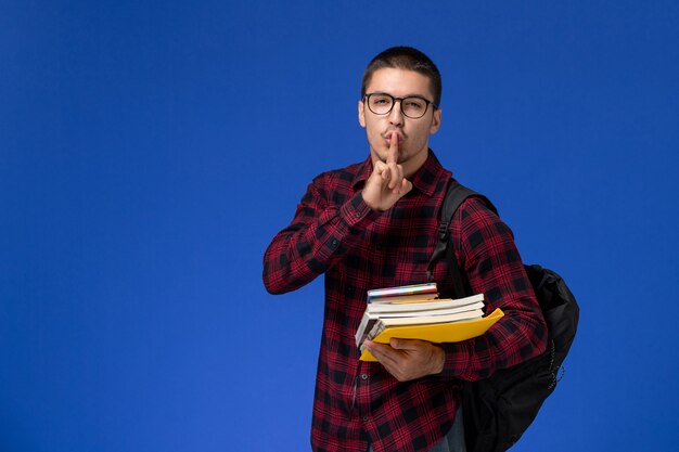 Front view of male student in red checkered shirt with backpack holding copybooks on light blue wall