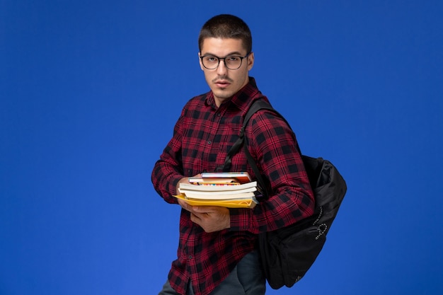 Front view of male student in red checkered shirt with backpack holding copybooks on light blue wall