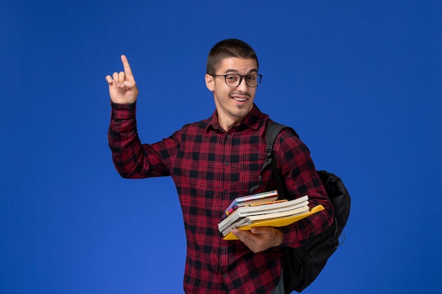 Front view of male student in red checkered shirt with backpack holding copybooks smiling on light-blue wall