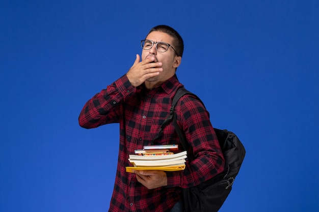 Front view of male student in red checkered shirt with backpack holding copybooks yawning on light-blue wall