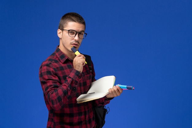 Вид спереди студента в красной клетчатой рубашке с рюкзаком, держащего тетрадь с фломастерами на голубой стене