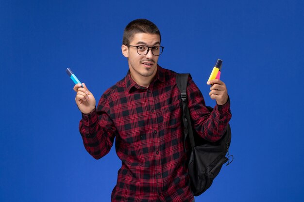 Вид спереди студента в красной клетчатой рубашке с рюкзаком, держащего фломастеры на голубой стене