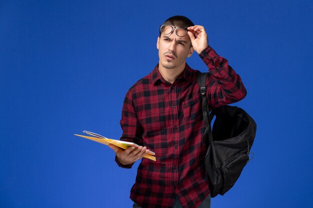 Вид спереди студента в красной клетчатой рубашке с рюкзаком с файлами и тетрадью на голубой стене