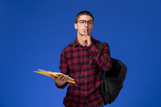 Front view of male student in red checkered shirt with backpack holding files and copybook on light blue wall