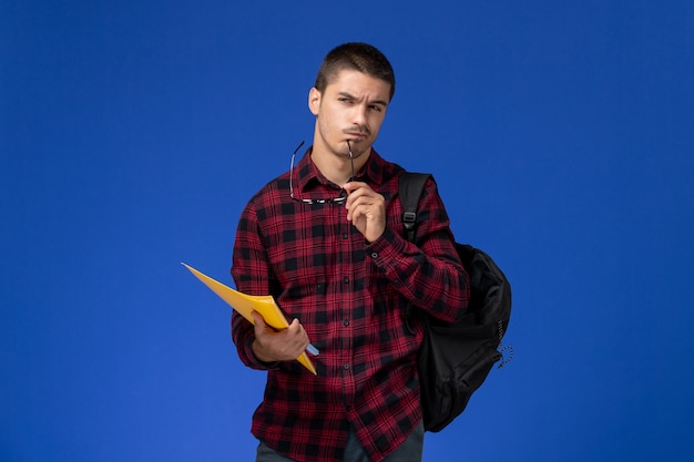 Front view of male student in red checkered shirt with backpack holding files and copybook thinking on light-blue wall