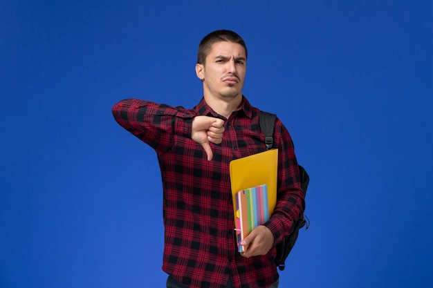 Foto gratuita vista frontale dello studente maschio in camicia a scacchi rossa con lo zaino che tiene file e quaderni sulla parete blu