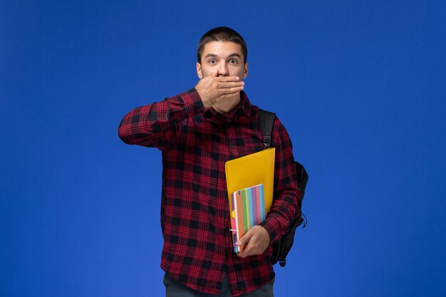 Вид спереди студента в красной клетчатой рубашке с рюкзаком, держащим файлы и тетради на синей стене