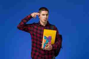 Free photo front view of male student in red checkered shirt with backpack holding files and copybooks on light-blue wall