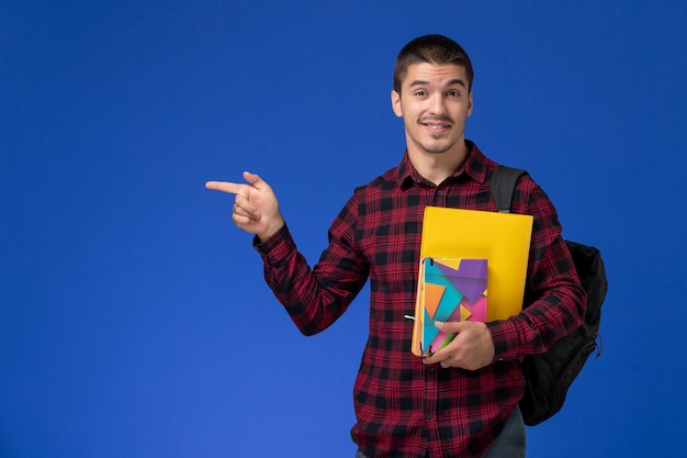 Front view of male student in red checkered shirt with backpack holding files and copybooks on light-blue wall