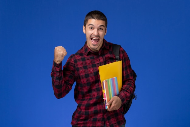 파란색 벽에 기쁨 파일과 카피 북을 들고 배낭과 빨간색 체크 무늬 셔츠에 남성 학생의 전면보기