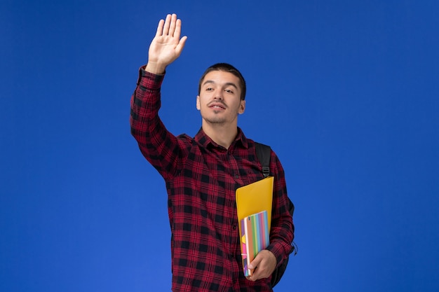 Foto gratuita vista frontale dell'allievo maschio in camicia a scacchi rossa con lo zaino che tiene file e quaderni agitando la mano sulla parete blu