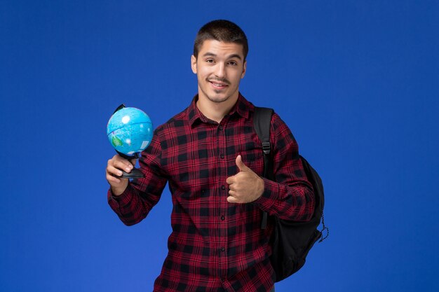 Front view of male student in red checkered shirt with backpack holding little globe on blue wall