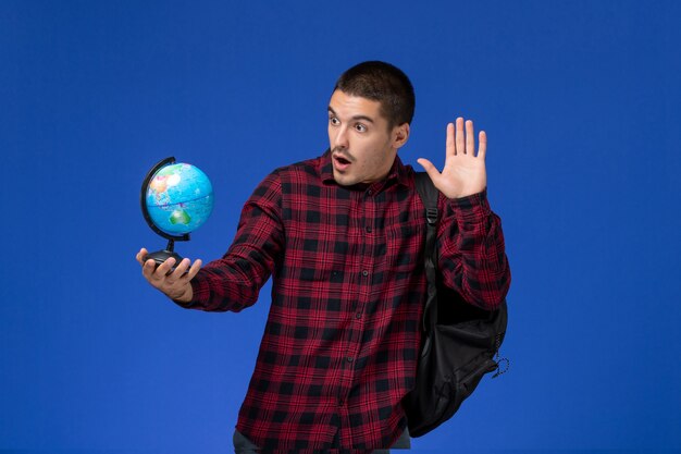 Front view of male student in red checkered shirt with backpack holding little globe on light-blue wall