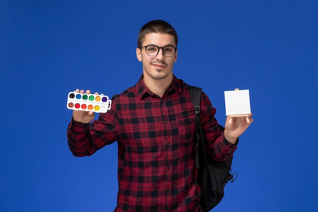 Front view of male student in red checkered shirt with backpack holding paint and easel on blue wall
