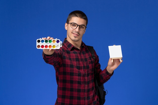 Foto gratuita vista frontale di uno studente maschio in camicia a scacchi rossa con lo zaino che tiene vernice e cavalletto sulla parete blu