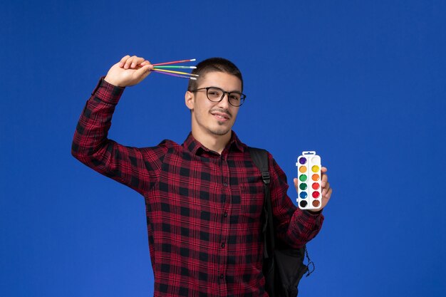 Вид спереди студента в красной клетчатой рубашке с рюкзаком, держащим краски для рисования и кисточки на синей стене