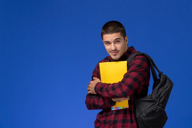 Front view of male student in red checkered shirt with backpack holding yellow files on the blue wall