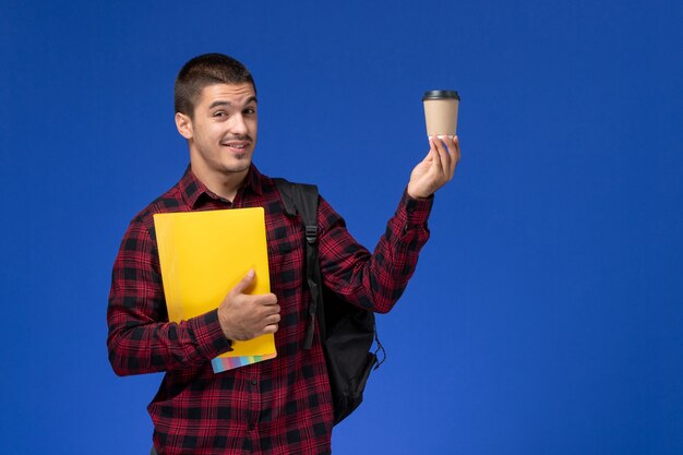 Front view of male student in red checkered shirt with backpack holding yellow files and coffee on the blue wall