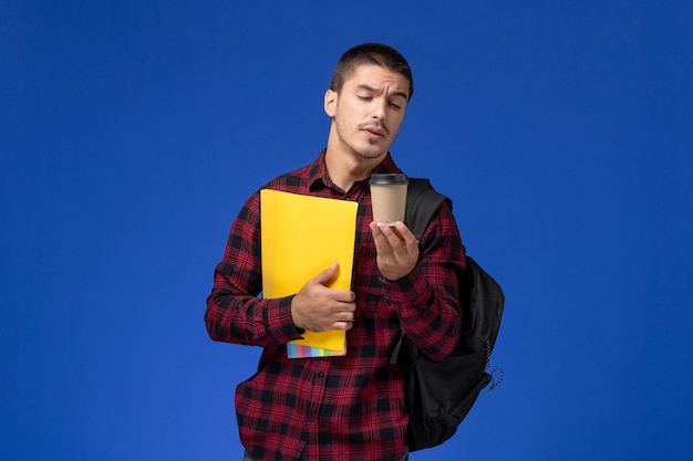 Front view of male student in red checkered shirt with backpack holding yellow files and coffee on the blue wall