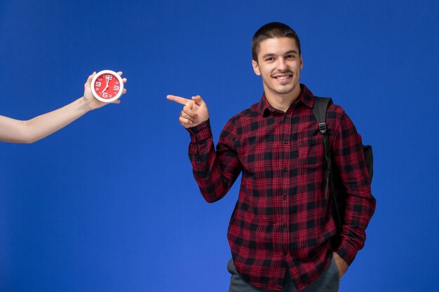 Front view of male student in red checkered shirt with backpack smiling on the blue wall