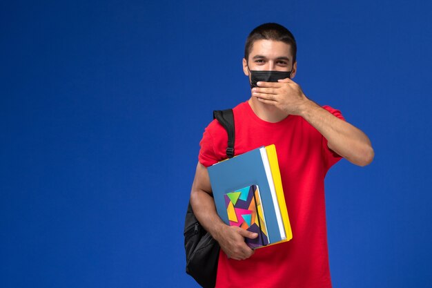 파란색 배경에 카피 북 및 파일을 들고 검은 살 균 마스크에 배낭을 착용하는 빨간 티셔츠에 전면보기 남성 학생.