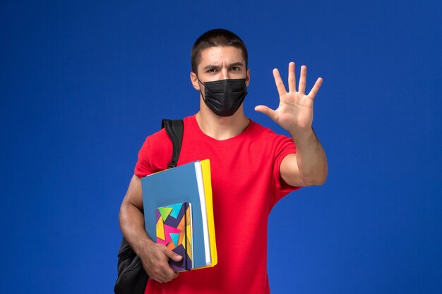 Front view male student in red t-shirt wearing backpack in black sterile mask holding copybook and files on blue desk.