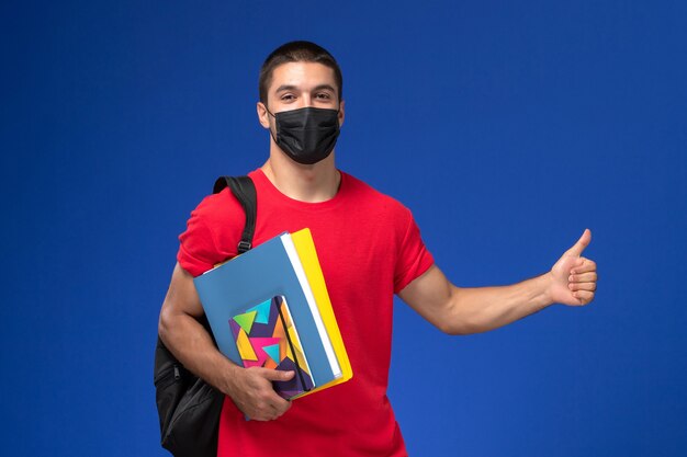 Front view male student in red t-shirt wearing backpack in black sterile mask holding copybooks on the blue background