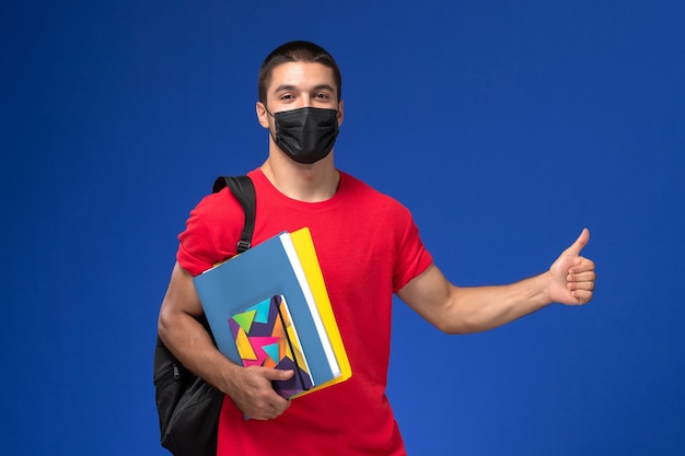 青い背景にコピーブックを保持している黒い滅菌マスクのバックパックを身に着けている赤いTシャツの正面図男子学生。
