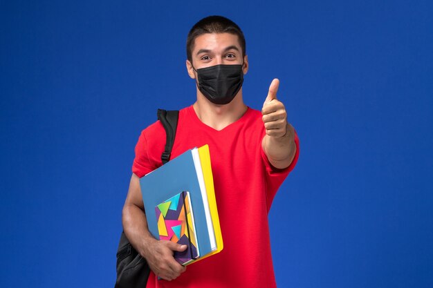 Front view male student in red t-shirt wearing backpack in black sterile mask holding copybooks on the blue background.