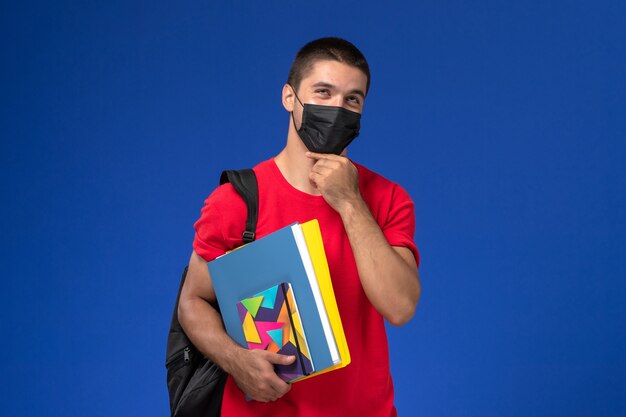 青い背景を考えてコピーブックを保持している黒い滅菌マスクのバックパックを身に着けている赤いTシャツの正面図男子学生。