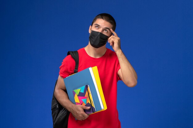 파란색 배경에 생각 카피 북을 들고 검은 살 균 마스크에 배낭을 착용하는 빨간 티셔츠에 전면보기 남성 학생.