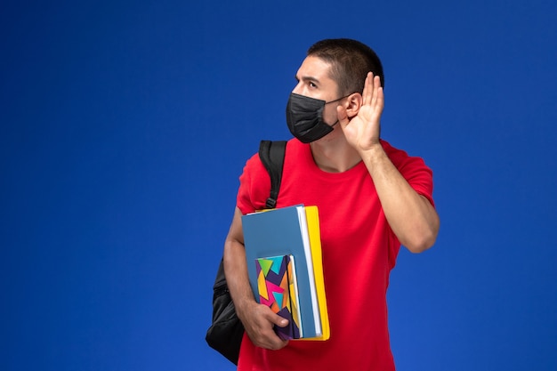 青い背景で聞くことを試みているコピーブックを保持している黒い滅菌マスクのバックパックを身に着けている赤いTシャツの正面図の男子生徒。