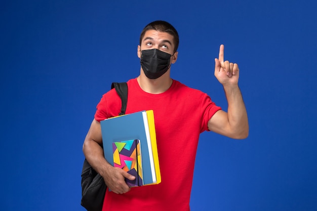 青い背景に上げられた指でファイルを保持している黒い滅菌マスクのバックパックを身に着けている赤いTシャツの正面図の男子生徒。
