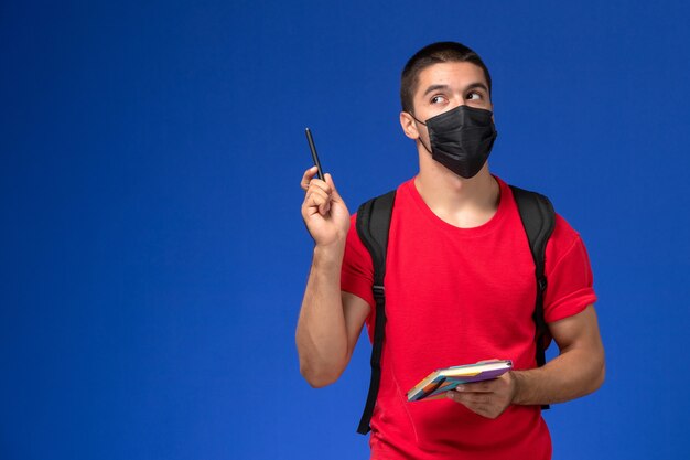 Студент вид спереди в красной футболке нося рюкзак в черной стерильной маске, держа ручку и тетрадь на синем фоне.