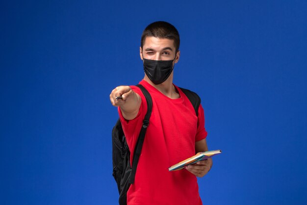 Front view male student in red t-shirt wearing backpack in black sterile mask holding pen and copybook on blue background.