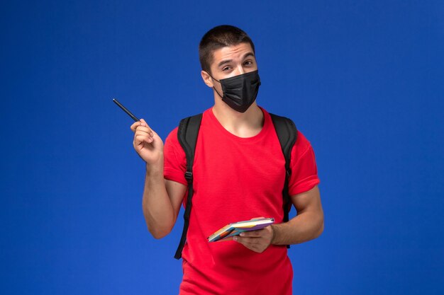 Front view male student in red t-shirt wearing backpack in black sterile mask holding pen and copybook on blue background.