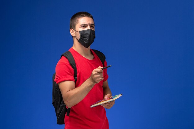 파란색 책상에 펜과 카피 북을 들고 검은 살 균 마스크에 배낭을 착용하는 빨간 티셔츠에 전면보기 남성 학생.