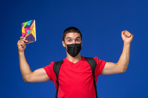 Front view male student in red t-shirt wearing backpack in black sterile mask holding pen and copybook rejoicing on blue background.