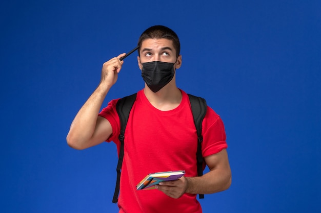 Front view male student in red t-shirt wearing backpack in black sterile mask holding pen and copybook thinking on blue background.